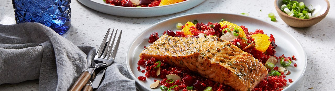 Two plates of grilled salmon with quinoa and orange slices on a white table with blue glasses, utensils, and a pan of more food in the background.